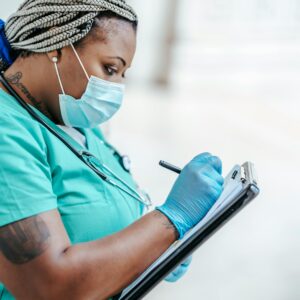 Side view of a healthcare professional in PPE writing on a clipboard indoors.