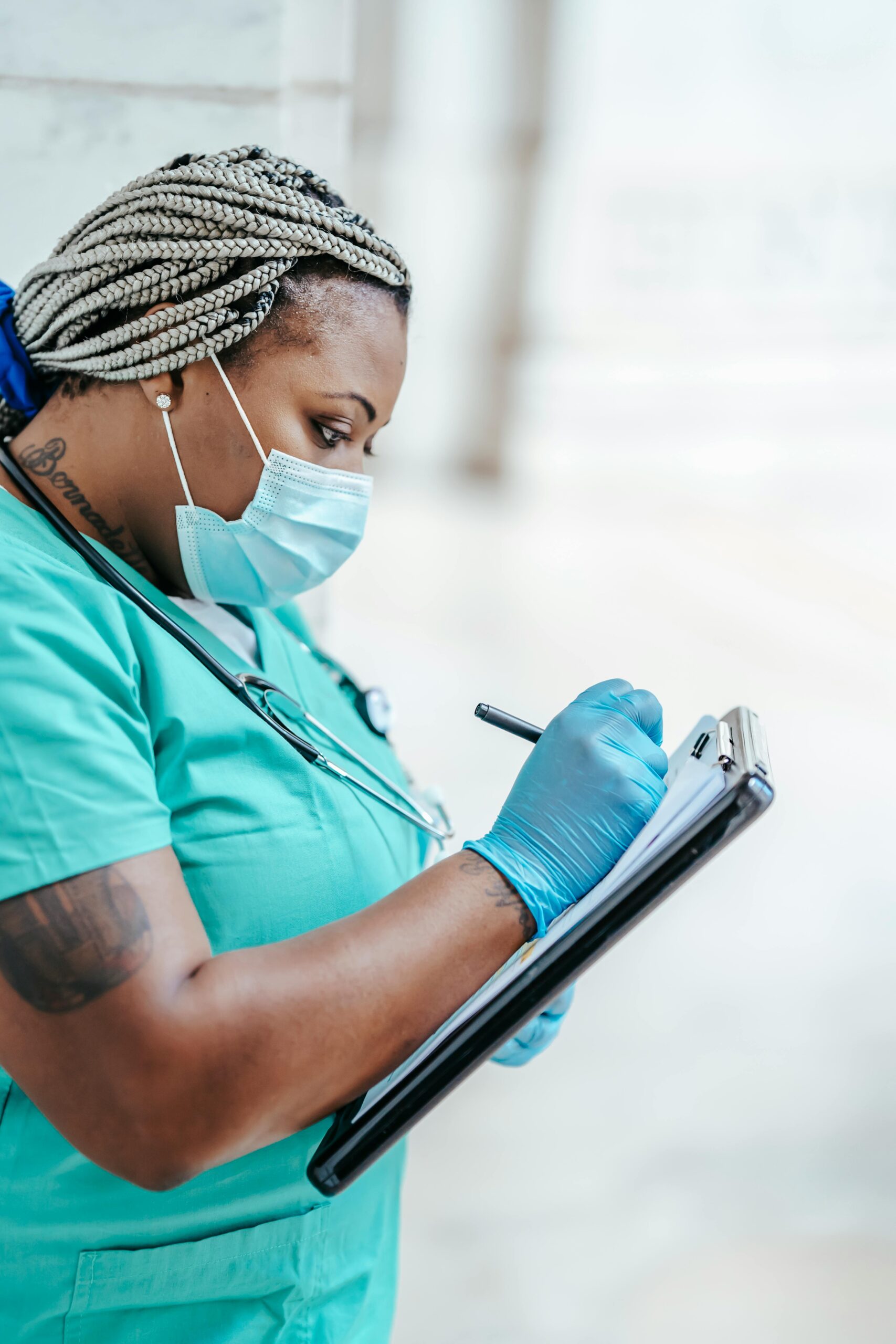 Side view of a healthcare professional in PPE writing on a clipboard indoors.